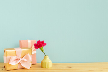 Gift boxes with vase of pink carnation flower on wooden table with blue background