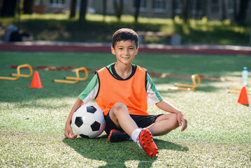 Happy little boy in uniform sitting on soccer field with a ball in front of him after training.