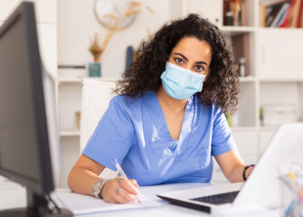 Fototapeta na wymiar Attentively young female doctor in protective mask working at laptop in her office