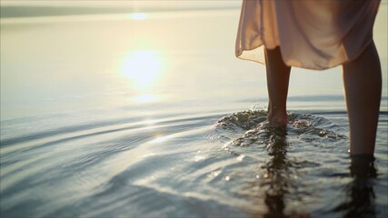 She is wearing a dress. Women's legs in a dress in the water of the lake at sunset. Details of woman's legs.
