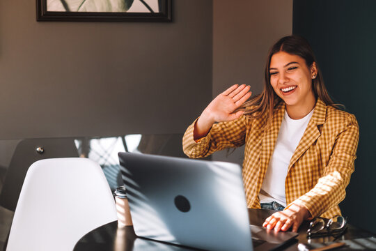 Portrait of happy stylish brunette young woman sitting at table, looking at laptop screen on video call and greeting with waving. Indoor studio shot, cafe, office background.