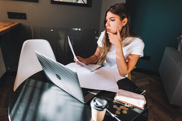 Pensive young caucasian woman calculating bills in kitchen, sitting at table with laptop computer,...
