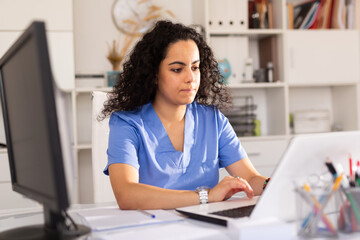 Young woman doctor assistant working in medical office using laptop computer and writing...