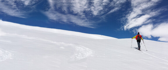 Naklejka na ściany i meble man ski touring climbing snow-covered mountain under blue and cloudy sky