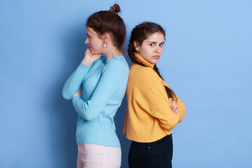 Two European girls going through conflict in their relationship, posing back to back isolated over blue background, keeping hands folded, wearing casual outfits.