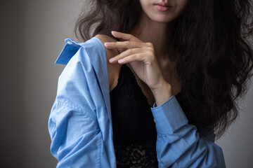 Portrait of young woman with brunette curly hair wearing blue oversize shirt and lacy bra over simple background.