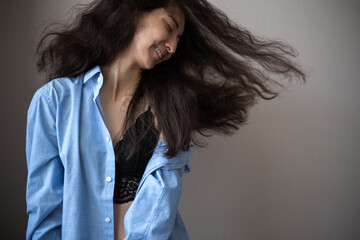 Portrait of young woman with brunette curly hair wearing blue oversize shirt and lacy bra over simple background.