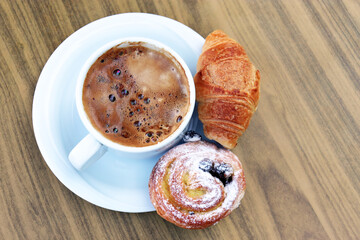 Cup of coffee and croissant and pastries in a white saucer on wooden table. Tasty breakfast, cappuccino and fresh cakes