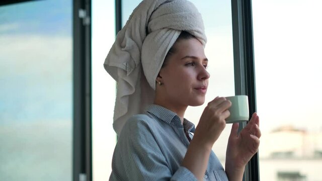 Woman with her hair wrapped in white towel enjoying her morning coffee or tea at the balcony. Concept. Portrait of a beautiful girl drinking hot beverage against an opened window.
