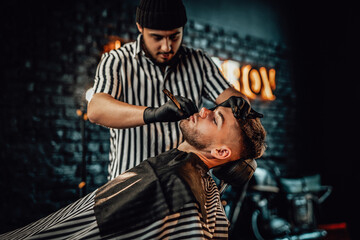 Fototapeta na wymiar Retro styled barbershop and man is shaving by caucasian barber with black gloves in blurred brickwall background.