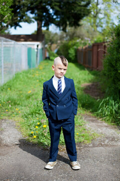 Boy in blue suit with mohawk stands in an overgrown alley, looking rather serious