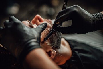 Skilled barber with modern sharp blade cutting his customer's beard in black gloves in his professional retro saloon.