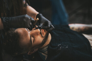 Guy sitting and relaxing in chair while he is shaving by skillful hairdresser with black gloves in...