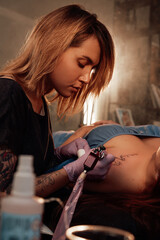 Pretty girl lying on bed and waiting while tattoo master finish drawing on her shoulder in warm and shiny saloon.