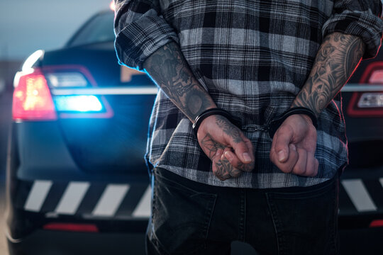 Tattooed arms & hands of a suspect in handcuffs behind a police cruiser
