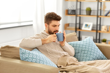 health, cold and people concept - sick young man in blanket drinking hot tea at home