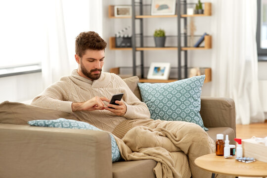 health, cold and people concept - sick young man in blanket with smartphone and medicine at home