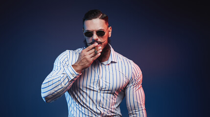 Obraz premium Strong and styled gentleman with beard and mustache posing holding a cigar in hand in dark background.