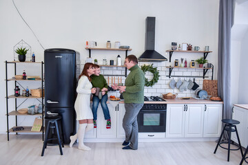 Happy family in the white kitchen in a Scandinavian style, drinking hot drinks and laughing. A young woman hugs her son, a man holds a gray cup in his hands. Minimalistic christmas wreath. Copy space 
