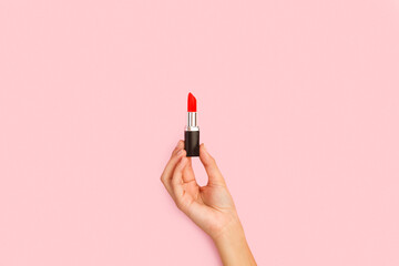 Mano femenina soteniendo un lápiz labial de color rojo sobre un fondo rosa pastel liso y aislado. Vista de frente y de cerca. Copy space. Concepto: Belleza - obrazy, fototapety, plakaty