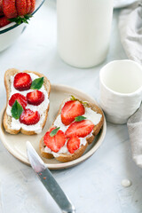 Toasts with cheese cream and slices of strawberries decorated with fresh mint leaves with a glass of milk on a white plate. Breakfast concept.