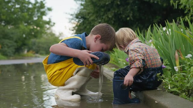 friendly childhood, attractive boy caring for his brother and takes off rubber boots pour out water while playing in puddle after rain in outdoor