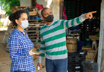 Fototapeta na wymiar Hispanic male and female farmers in protective face masks standing outdoors on farm discussing work topics. Pandemic precautionary concept