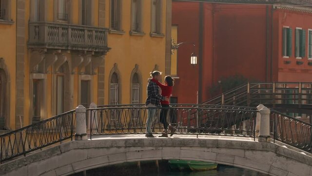 Handsome loving man kissing and hugging his charming asian girlfriend standing on the ancient Venetian bridge during their honeymoon. Travelling to Europe as the best way of spending the vacation.