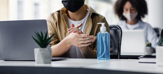 Focus on antiseptic to protect against covid virus. Guy disinfects his hands, sitting at table at workplace with laptop, working in office