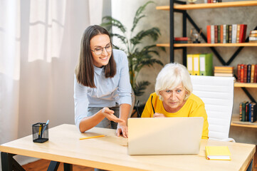 A young female coworker and older female coworker look on laptop screen and discuss some business tasks in the contemporary office. Concept of diverse generations in the team