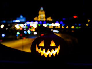 Halloween pumpkin on the background of the night city. Multi-storey buildings and skyscrapers. Blurry colored lights of city illumination. Night city decor with a festive Halloween theme. Copy space.