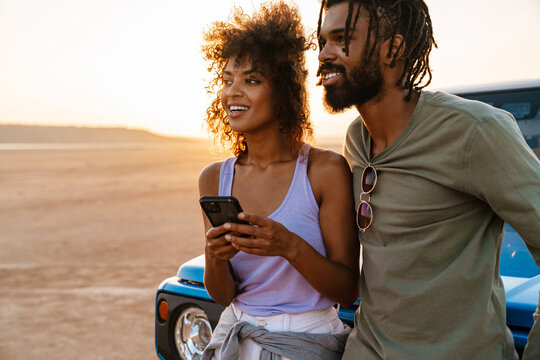 Image of joyful african american couple using mobile phone while travelling with car