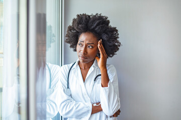 Doctors have many stresses to deal with too. African American female doctor with headache. Young female healthcare worker having an headache and touching her head in hospital lobby