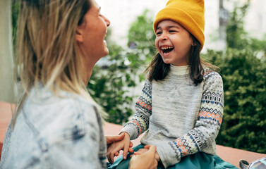 Side view image of cheerful little girl in red cap hugging her mom in yellow cap. Cute kid embracing her mother enjoying the time together outside. Mother and daughter share love. Mother's day.