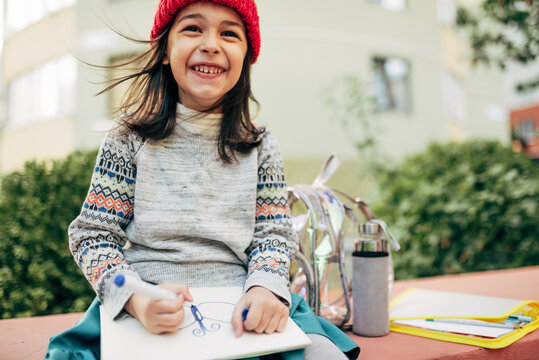 Horizontal Image Of Cute Smiling Little Girl In Red Cap Is Sitting Outside And Drawing On Paper.  Pretty Kid Doing Homework Outside After The School.