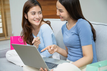 Beautiful young asian women happy couple sitting on sofa buying online using laptop a computer and phone in living room at home.