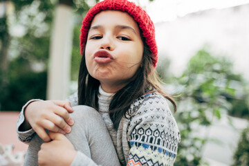 The outdoor portrait of a playful little girl in a red winter hat is sitting outside and blowing a kiss to the camera. Pretty kid takes a break outside after school and making a grimace.