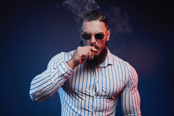 Brutal bearded guy with muscular body and sunglasses smoking cigar and posing in dark background.