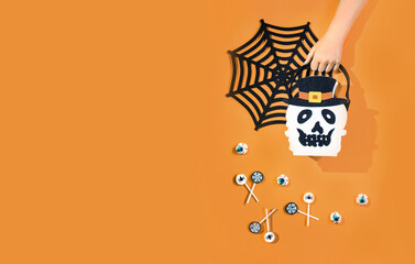 Child hand holding Halloween candy bucket face  bag of Jack O'Lantern with lollypops with spiders and nets on orange background and bloody eyes sweets. Flat lay trick or treat with copy space