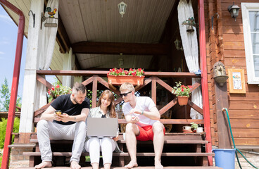 Young positive people discussing business project, startup idea, creative work team work remotely in country house, remote workers sitting outdoors on terrace. Three friends look on laptop screen