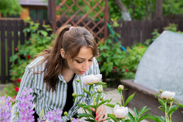 Fototapeta premium Attractive young woman gardener smelling fragrant white flowers of blooming peony in outdoor flower bed with eyes closed and smile, home garden, summer spring time, beauty, aroma, happy life concept