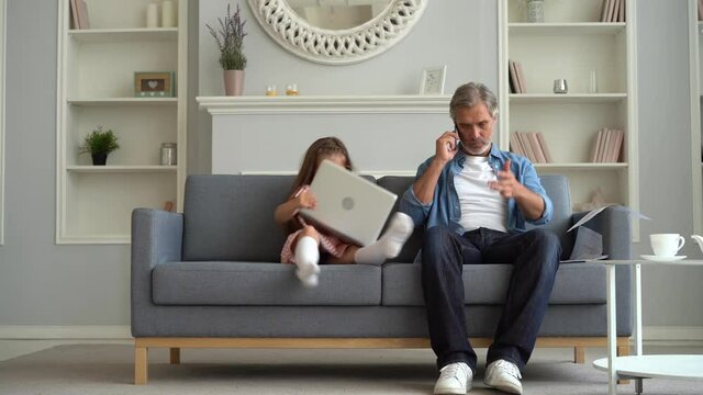 Busy caucasian father using laptop for distant work with daughter in living room. Upset child wants to play with dad