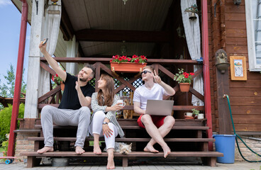 Young positive friends chilling on summer vacation, creative work team work remotely in country house, two men and woman making selfie sitting outdoors on terrace. Digital, gadgets concept, lifestyle