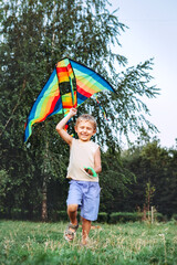 Cheerful little boy running with a multicolored kite on the city park green grass meadow. Funny childhood concept image.