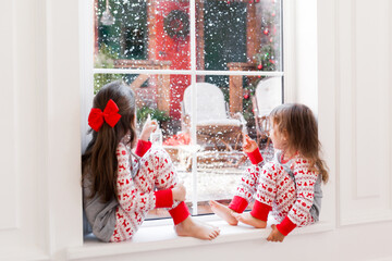 Two cute girls in pajamas sitting and looking out the window at snowy weather. Christmas moments...