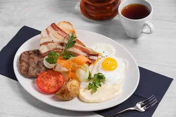 Breakfast of grilled beef cutlet, crispy bacon with ciabatta, fried eggs, grilled and fresh vegetables and cream cheese, cap of tea are on a white plate and a light background. Concept healthy food