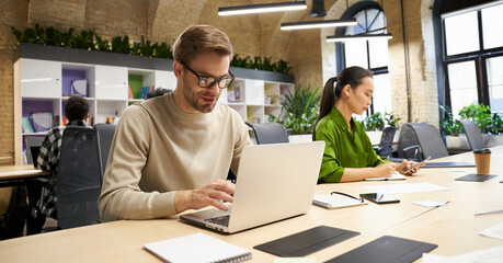 Young focused caucasian man wearing eyeglasses working on laptop while sitting at desk with his female colleague in the modern coworking space