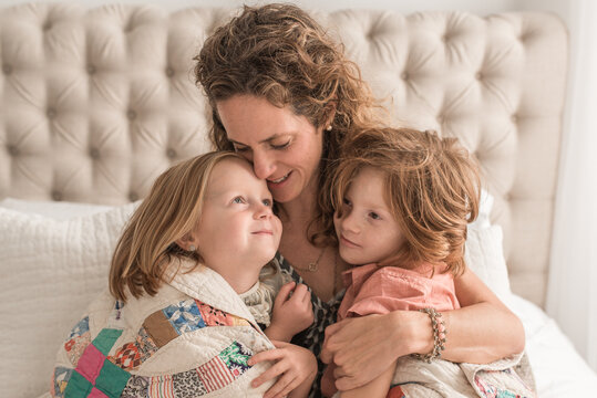 A Curly Haired Mother Snuggles With Her Children Under A Family Quilt