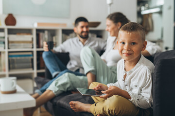 portrait of boy sitting on the sofa and using tablet computer, parents in blurred background
