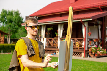 Young handsome artist in yellow sunglasses painting on easel outside on loan near country house, outdoors relaxing activity, creative hobby, creation, inspiration, summer weekend recreation concept